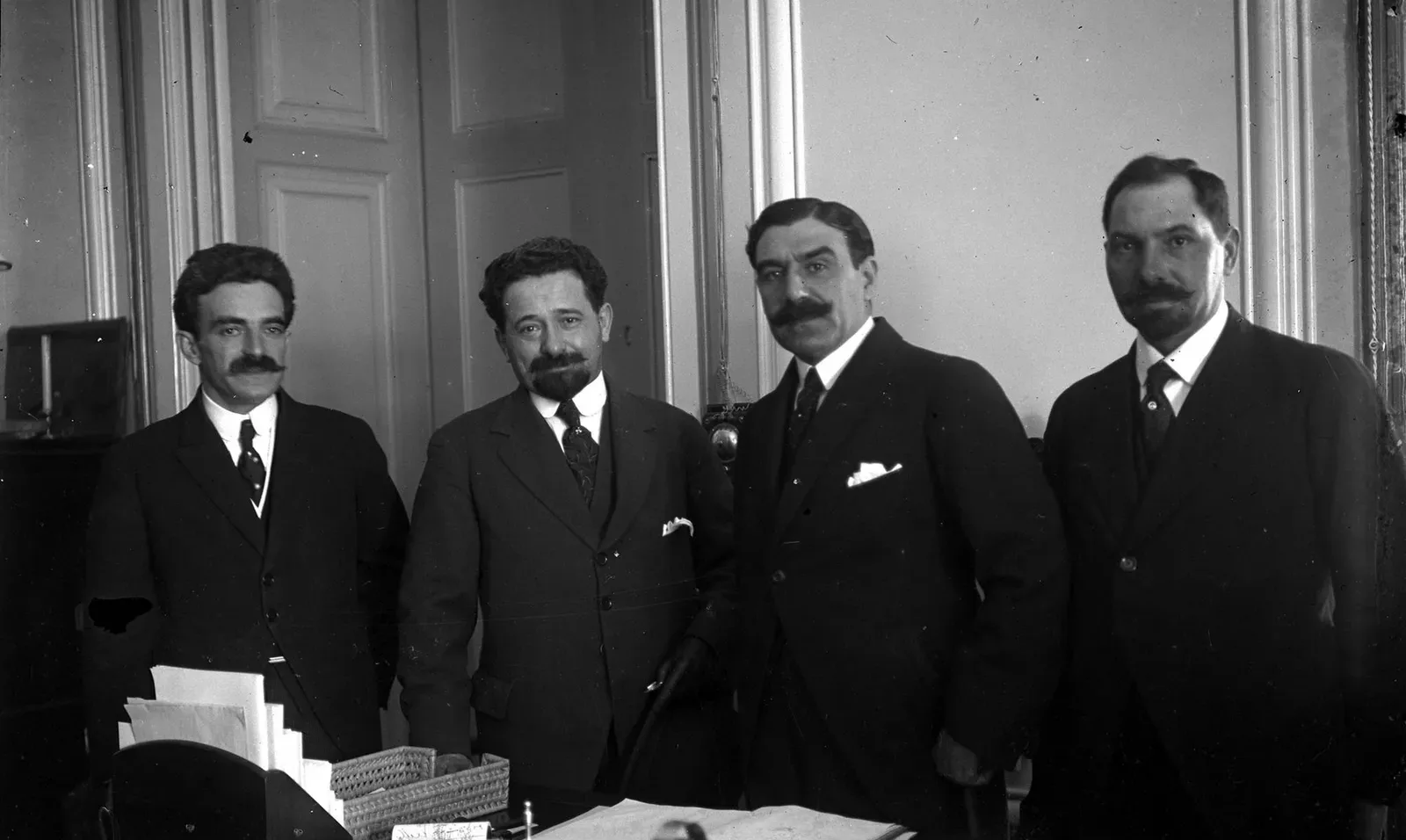 Four men in dark suits stand in a row behind a desk with papers, an inkstand and a letter tray, in a room with tall panelled doors. Three have heavy moustaches; the second from the left, shorter and bearded, looks straight at the camera.