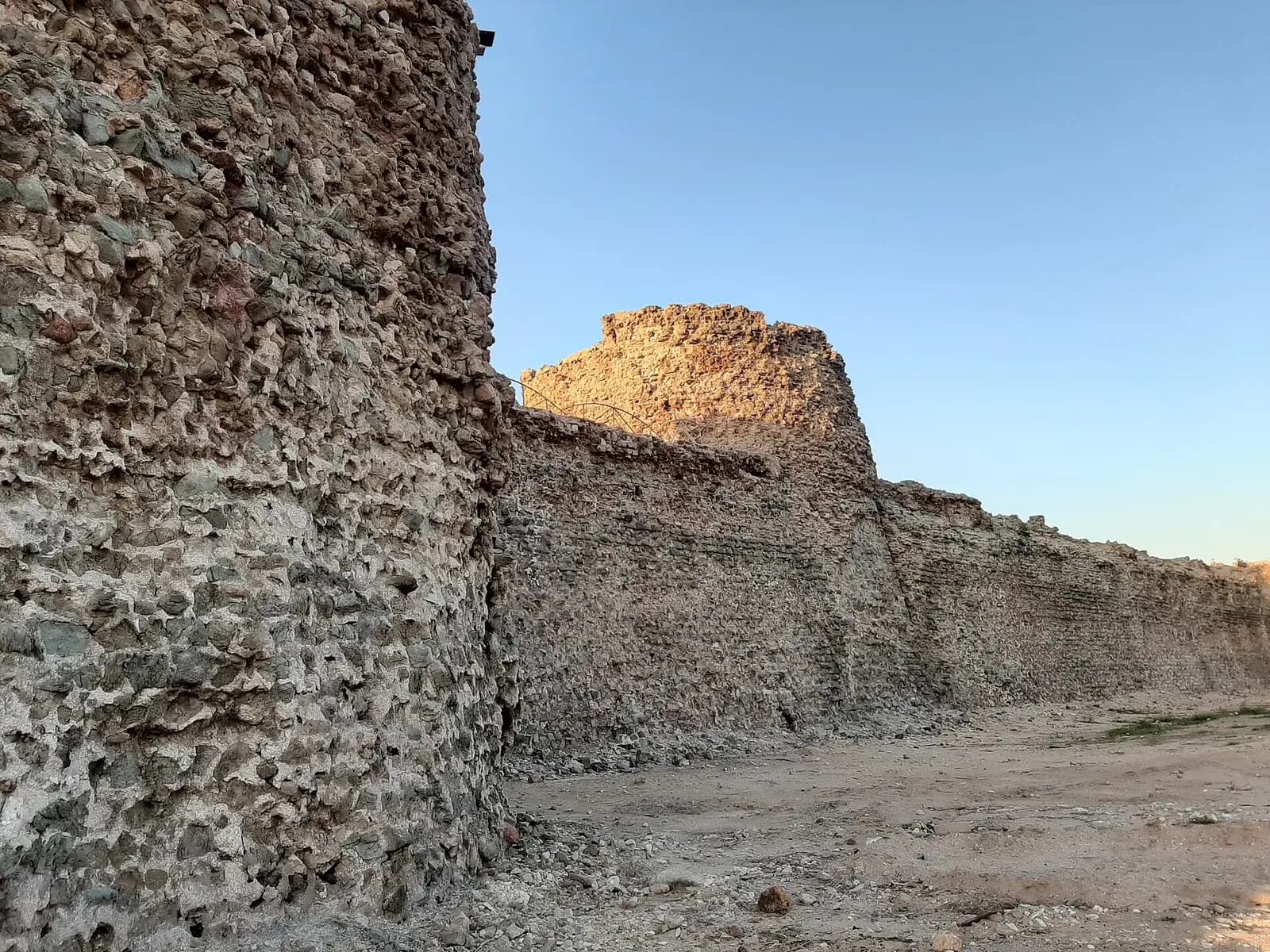 A massive rounded bastion of rough stones set in mortar fills the left of the picture. From it a high, weathered wall runs away from the camera to a ruined tower lit by the low sun, under a clear blue sky. Loose rubble lies on the ground at its foot.