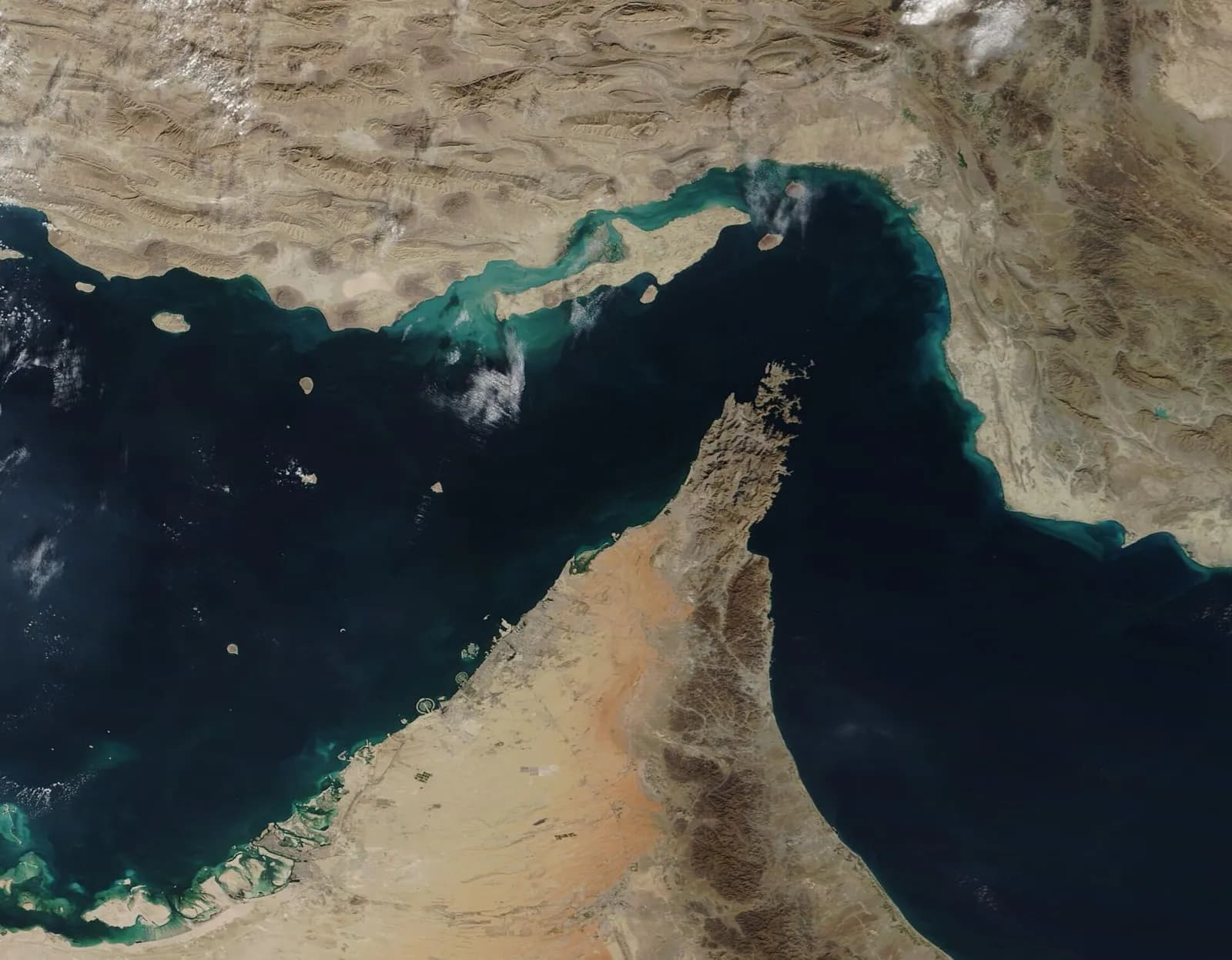 Satellite photograph of a narrow, dark blue sea passage. Brown, folded mountains of Iran fill the top; a long island lies along the Iranian shore with a small round island to its east. A rugged finger of land points up from the bottom between two wide stretches of sea.