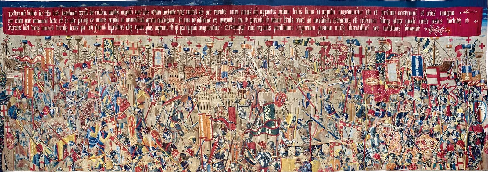 A crowded woven battle scene: hundreds of armoured soldiers with lances, banners and ladders swarm around the walls and towers of a town, under a long red band of Gothic Latin lettering.