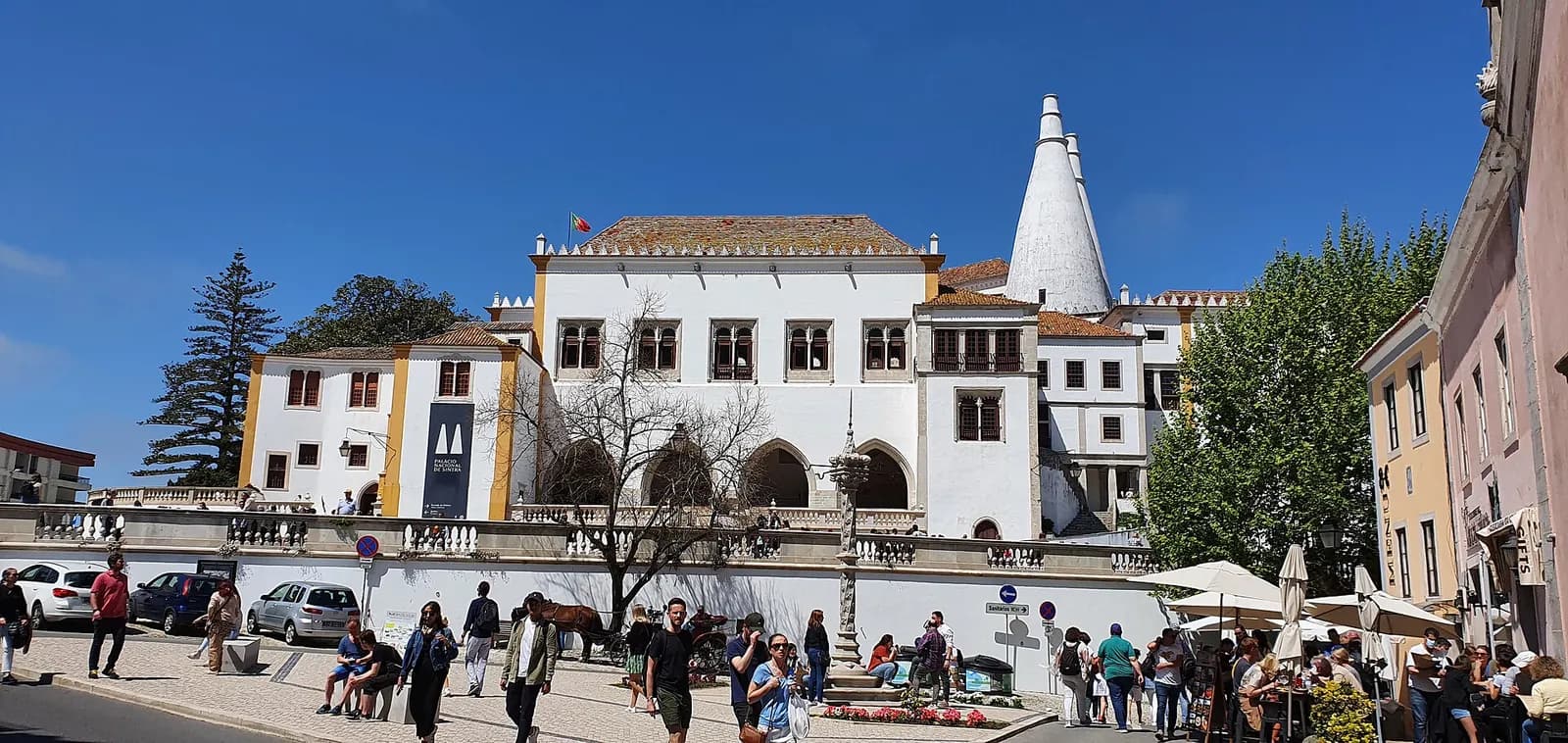 A long white palace with arched windows and two tall white conical chimneys stands behind a stone terrace, under a clear blue sky, with people walking in the square in front.