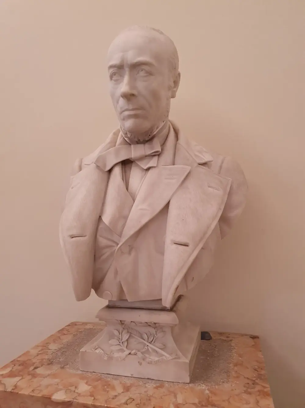 A white bust of a stern, clean-shaven man with side whiskers, a bow tie and a buttoned coat, on a pedestal carved with a laurel wreath, a quill and a ribbon reading História de Portugal, standing on a pink marble plinth.