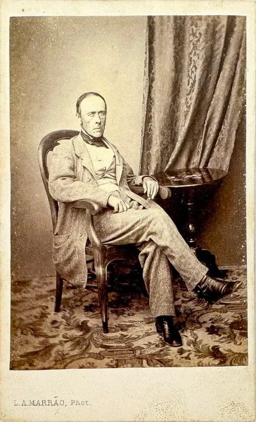 A carte-de-visite photograph of a balding, clean-shaven man with side whiskers, in a light tweed suit and dark cravat, sitting in a wooden armchair with his legs crossed beside a small table and a heavy curtain.