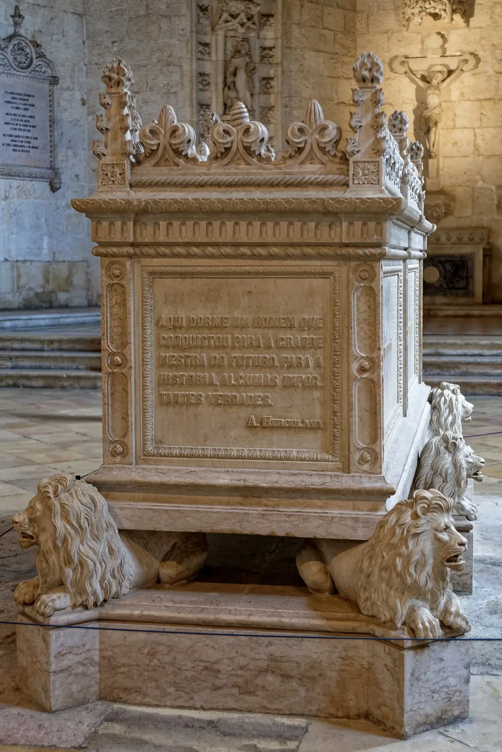 A tall carved stone tomb chest resting on crouching lions, its top crowned with Gothic-style pinnacles and leafy crests. A panel on the end bears a carved inscription signed A. Herculano. Behind it are stone walls, a crucifix and a carved niche.