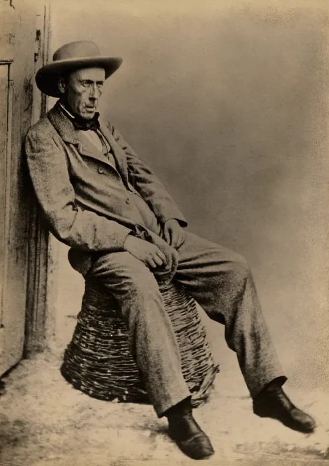 An old photograph of a thin older man in a wide-brimmed hat, rumpled tweed jacket and trousers and heavy boots, sitting on an upturned wicker basket and leaning back against a door frame.
