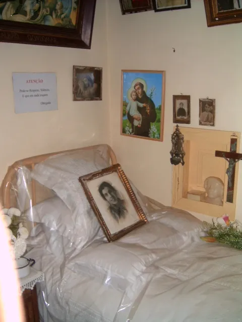 A narrow bed with white pillows under a clear plastic cover, with a framed charcoal portrait of a young woman propped on it. Religious pictures, a small crucifix and a printed notice hang on the cream walls.