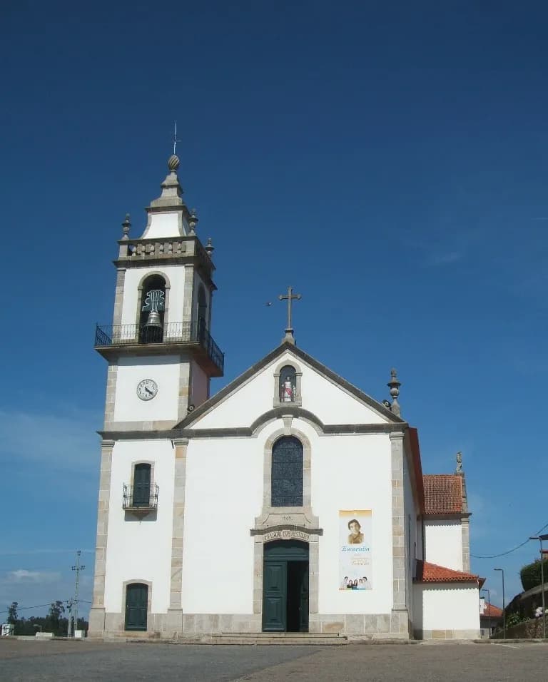 A whitewashed church with granite trim and a tall bell tower on the left, under a deep blue sky. A banner with a woman's portrait hangs beside the green front door.