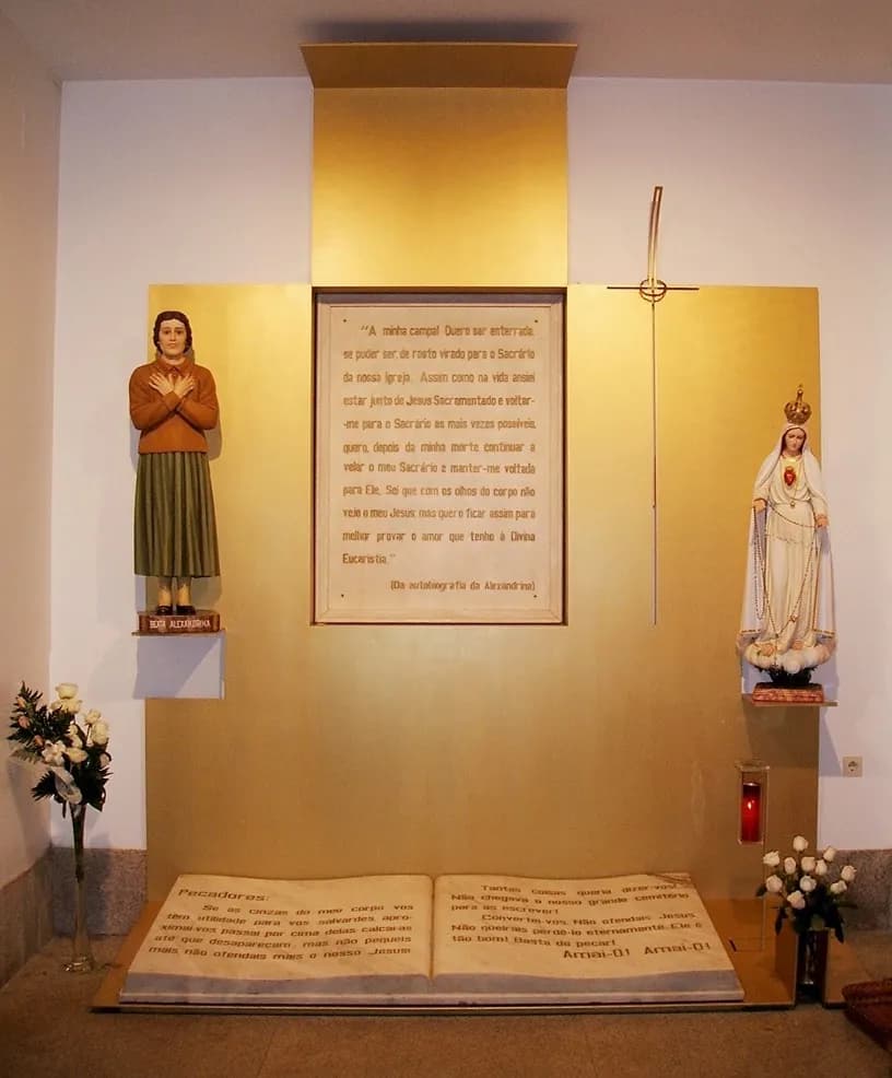 A gilded wall panel with a framed text in the centre, a painted statue of a young woman in a brown cardigan on the left and a statue of Our Lady of Fátima on the right. On the floor below lies a large open book carved in stone, inscribed with text.