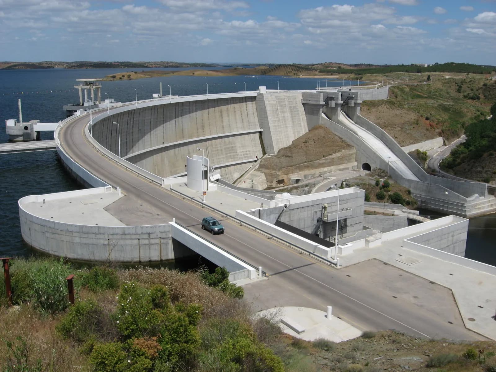 A curved concrete dam wall holds back a wide blue reservoir. A road with a single car runs along the top of the dam, and a concrete spillway chute runs down a scrubby hillside to the right.