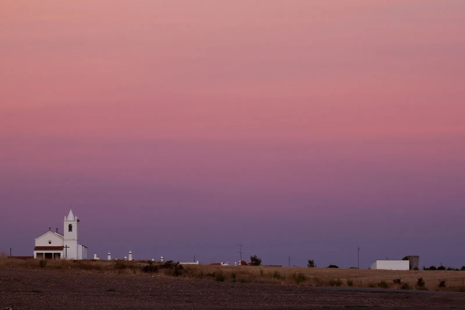 Under a pink and violet dusk sky, a small white church with a pointed bell tower stands at the left edge of a flat, dry field, with a few low white rooftops and chimneys strung out along the horizon.