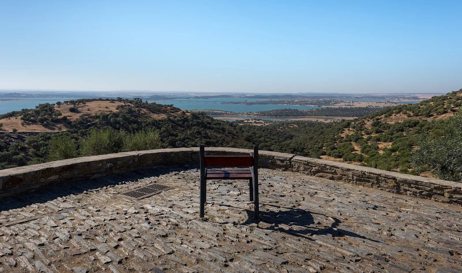 From a slate-paved terrace edged by a low stone wall with a single bench, a view across green, tree-dotted hills to a wide blue lake scattered with islands and crossed by a long bridge, under a pale sky.