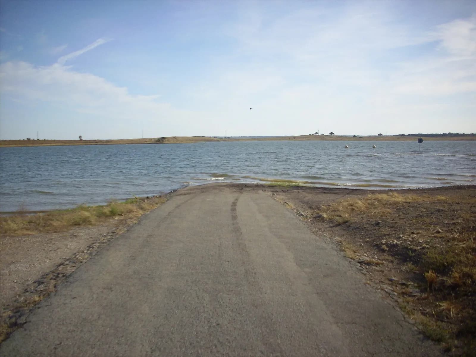 A narrow tarmac road runs straight ahead and disappears into the grey-blue water of a wide lake, with low, bare hills on the far shore.