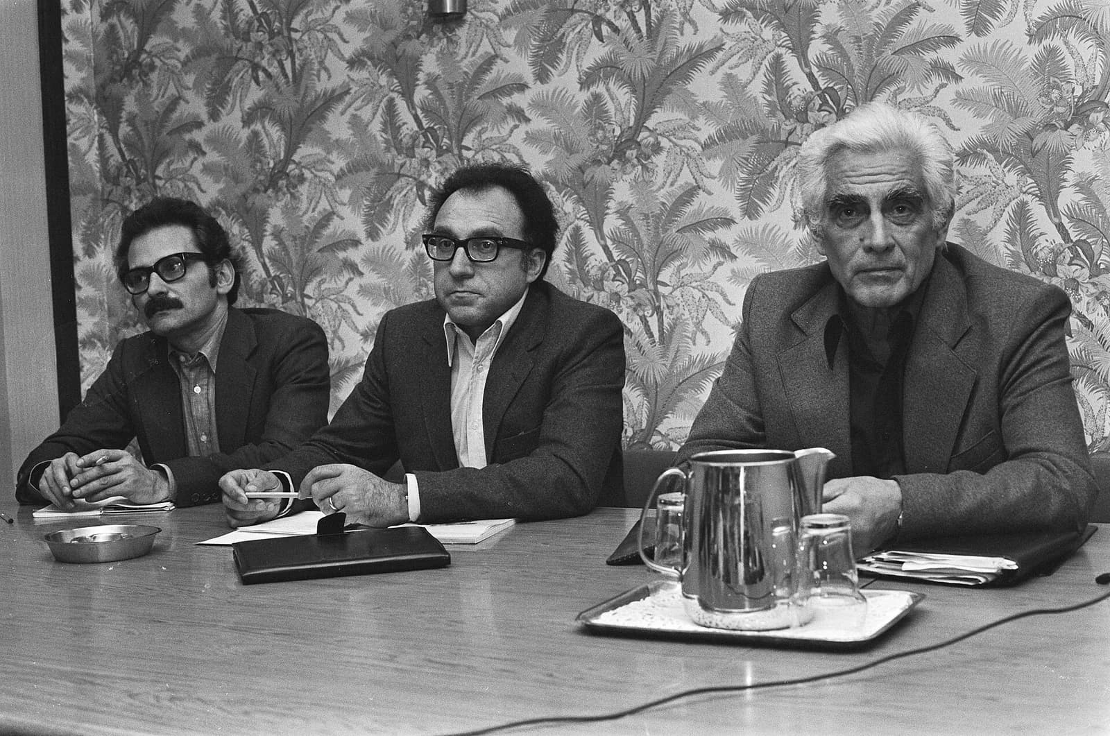 Black-and-white photograph of three men seated at a table in front of patterned palm-leaf wallpaper. The white-haired man on the right, in a dark jacket and open-necked shirt, looks at the camera; a water jug and glasses stand on a tray in front of him.