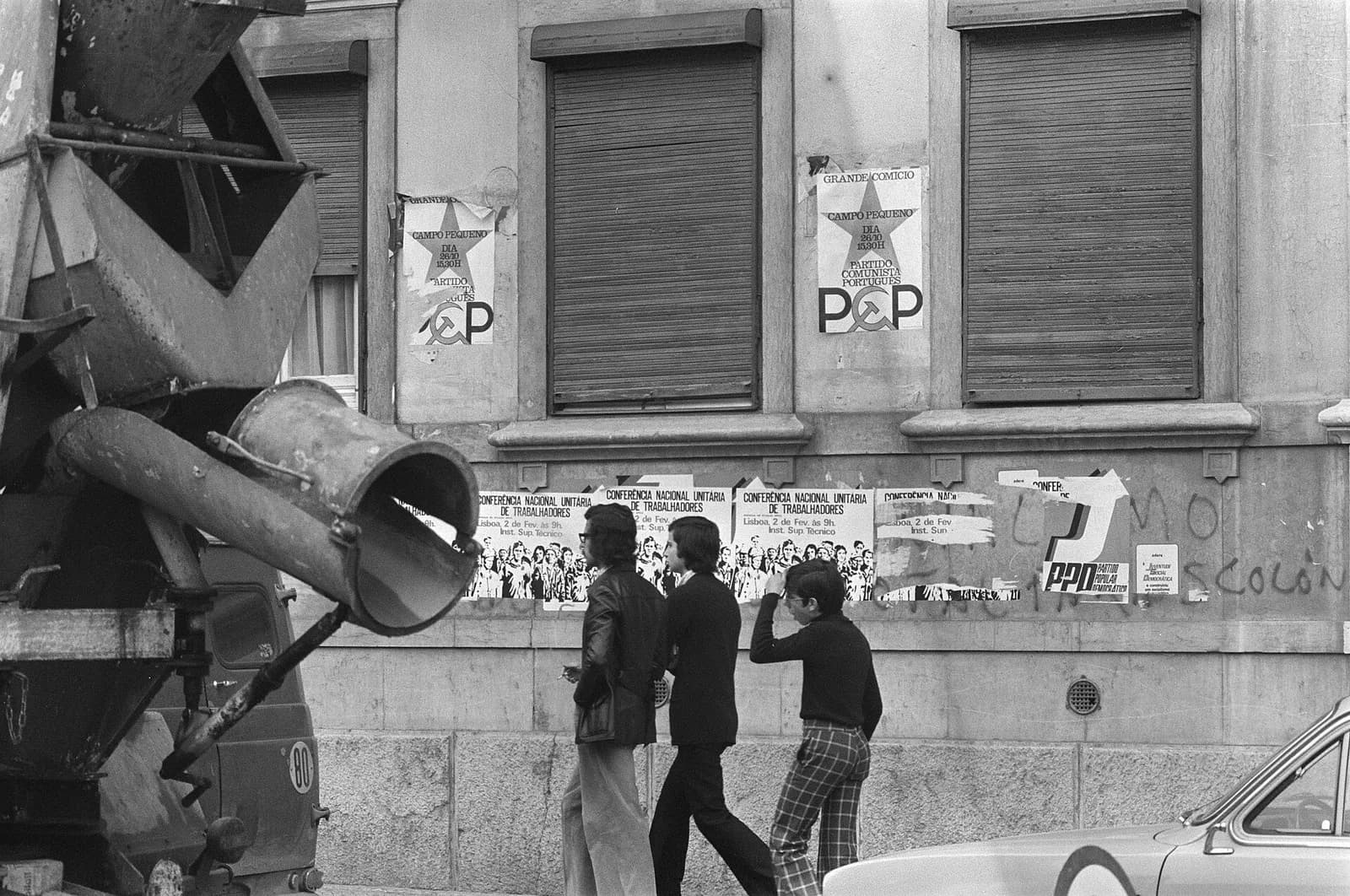 Black-and-white street scene: three young men walk past a building with shuttered windows, with a cement mixer in the foreground. Posters on the wall advertise a PCP rally at Campo Pequeno, a workers' conference and the PPD.