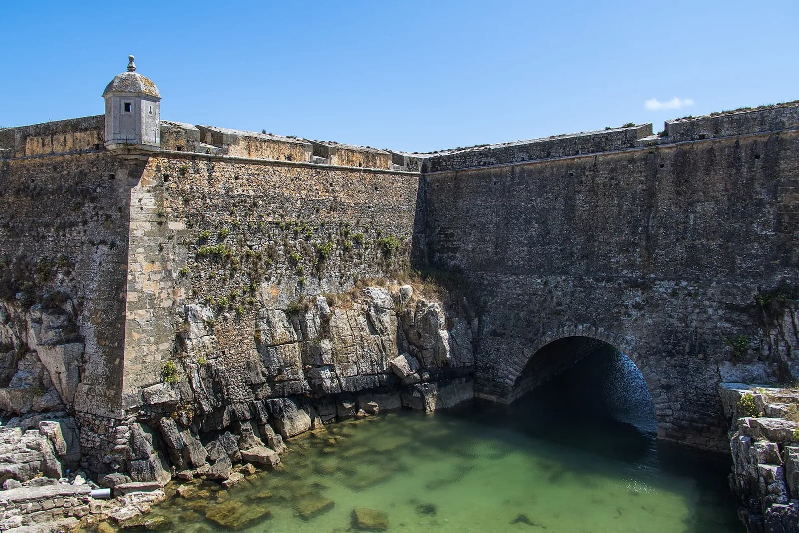 A tall wall of weathered grey stone rises straight out of rocks and clear green seawater, with a small domed sentry box on its corner and a stone arch at water level.