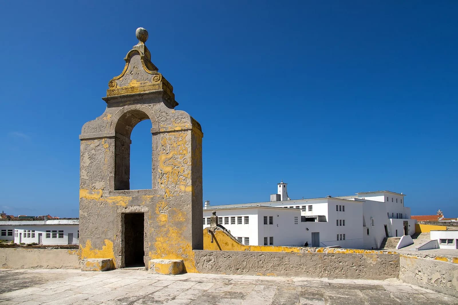 On a sunlit stone terrace stands an old arched sentry turret stained with yellow lichen. Behind it rise white, flat-roofed buildings of three storeys with rows of small windows.