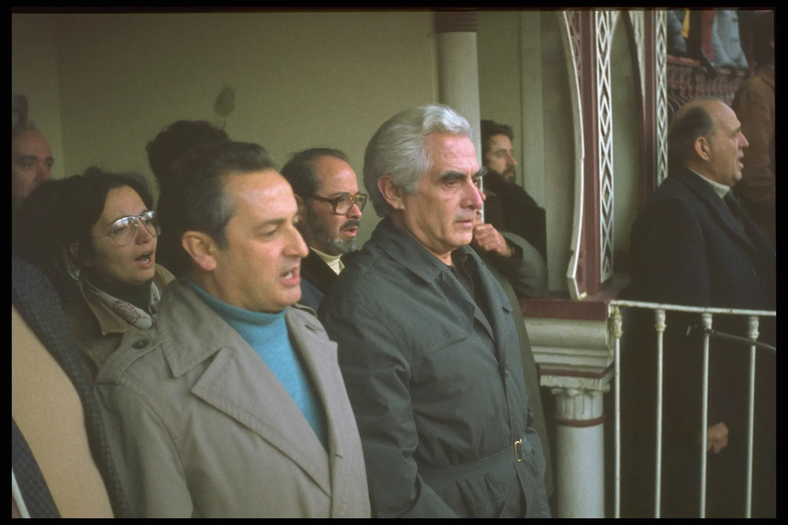 A grey-haired man in a dark green raincoat stands among other people at a balcony rail inside an arena with ornate red and white ironwork. Beside him a man in a beige coat and turquoise jumper is singing, and a woman in large glasses behind them is singing too.