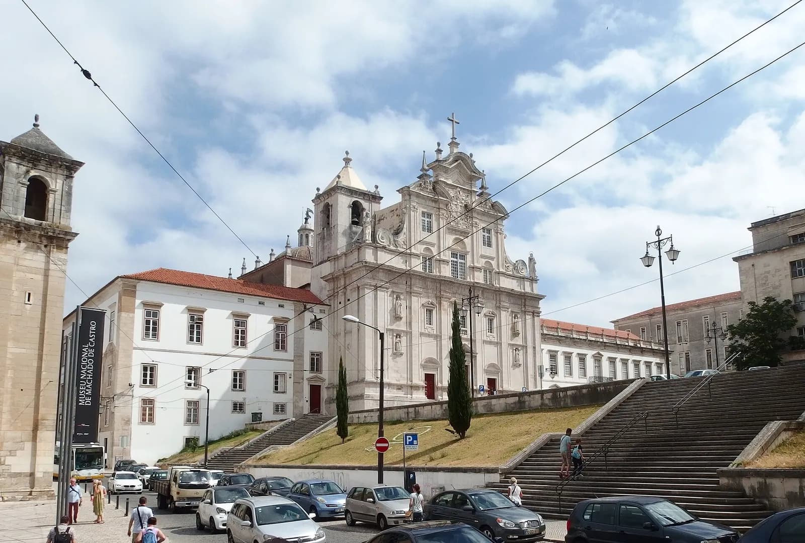 The pale stone baroque facade of a large church, with a bell tower on its left, stands above a grassy bank and wide flights of stone steps, with parked cars and pedestrians in the street below.