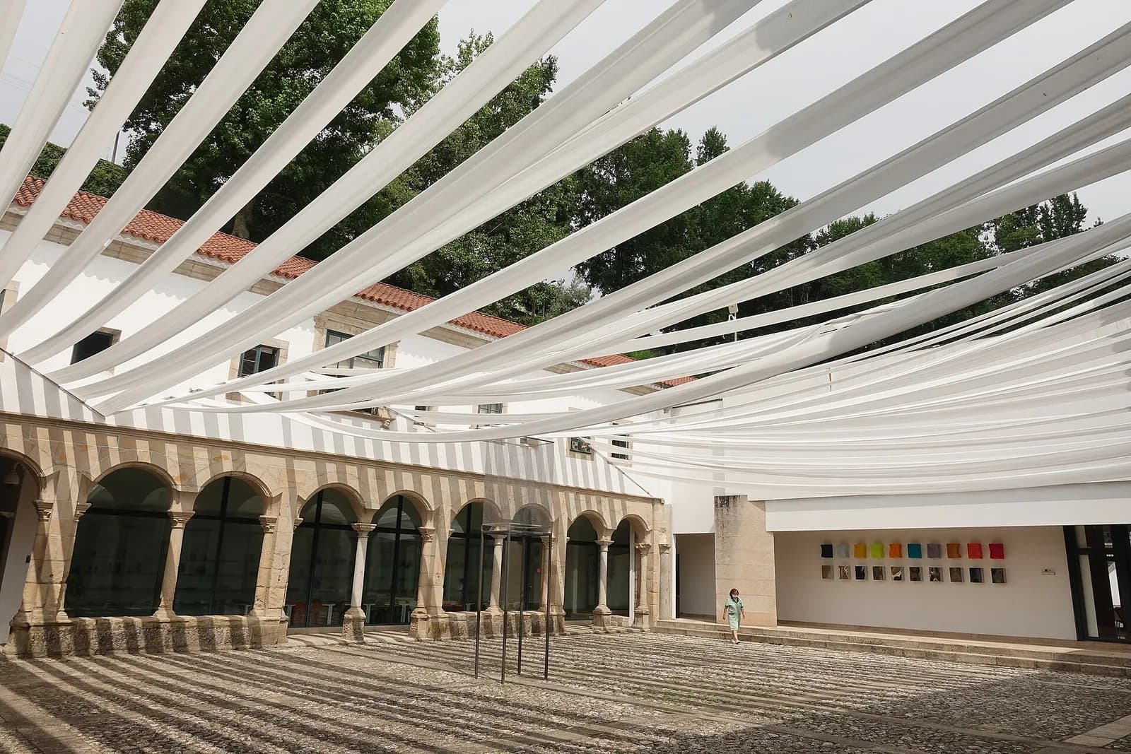 A cobbled courtyard shaded by long strips of white fabric stretched overhead. On the left is a two-storey whitewashed building with an arcade of stone arches on the ground floor; on the right a plain modern white wing with a row of small coloured panels on its wall. A visitor stands near the far corner.
