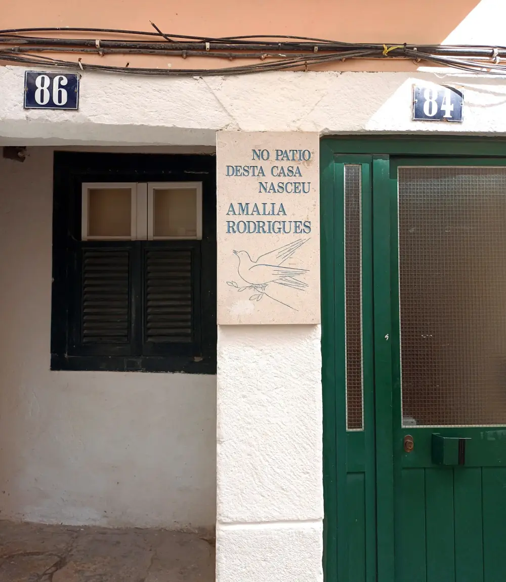 A whitewashed house front with blue number plates 86 and 84, a dark shuttered window on the left and a green door on the right. Between them a stone plaque engraved with a dove holding an olive branch reads No patio desta casa nasceu Amalia Rodrigues.