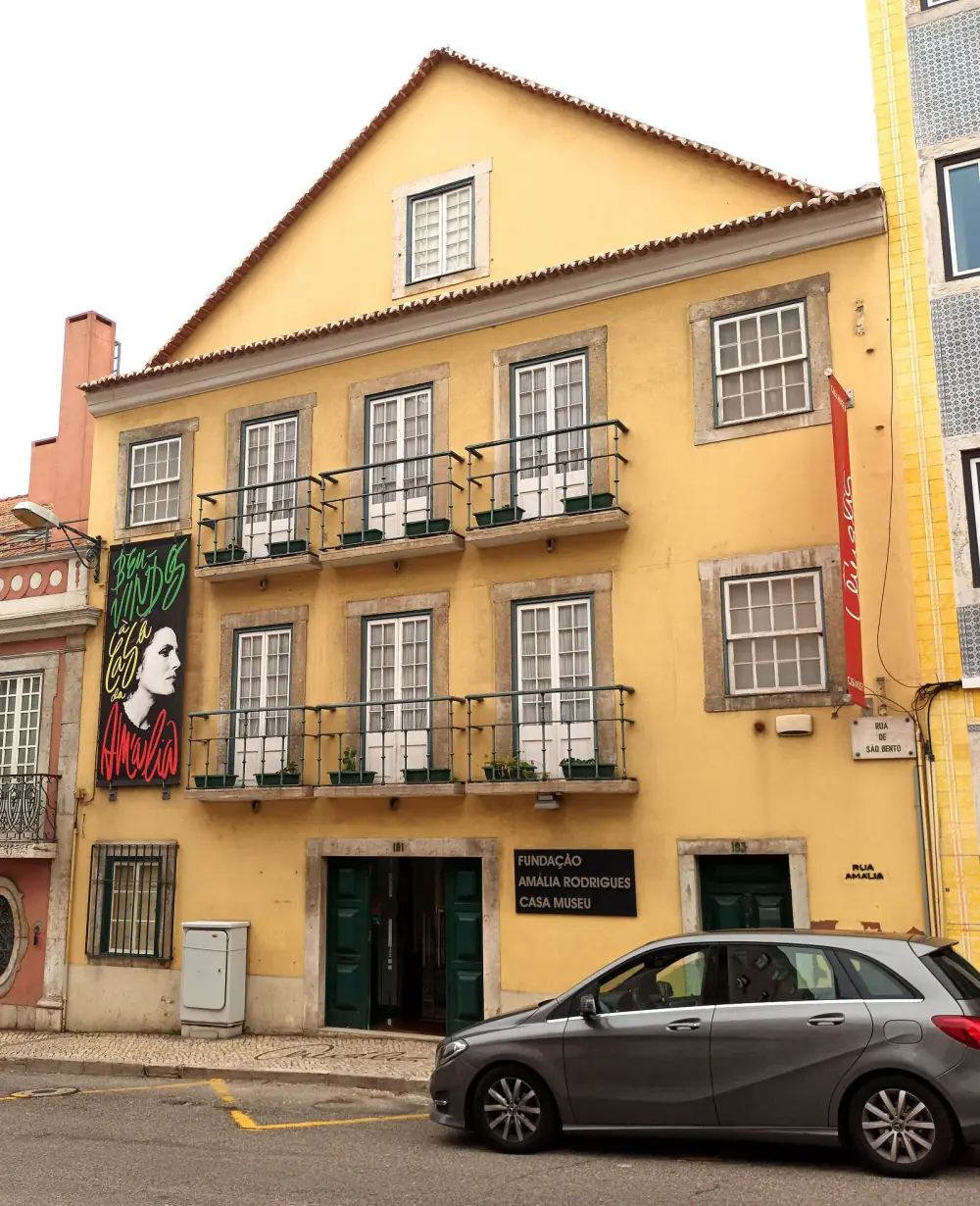 A three-storey yellow townhouse with iron balconies and a tiled roof. A sign by the green door reads Fundação Amália Rodrigues Casa Museu, and a banner with her portrait hangs on the left.