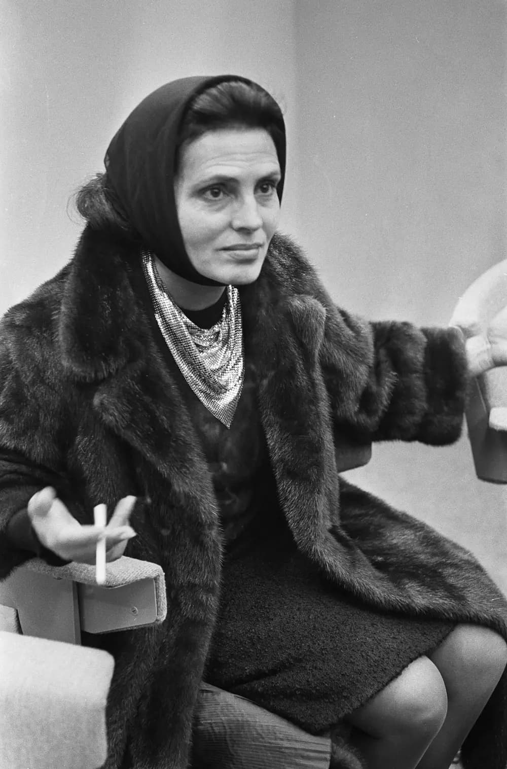 A woman in a dark headscarf, fur coat and a silver mesh scarf sits in an armchair holding a cigarette, looking to one side.