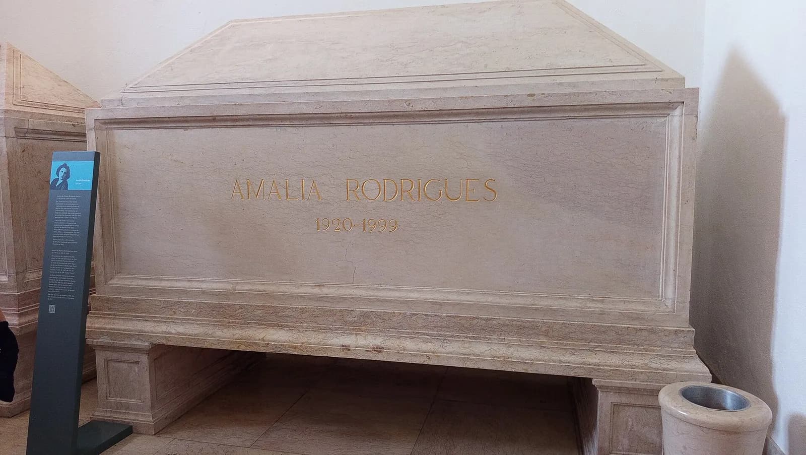 A large plain tomb of pale stone with a gabled lid, inscribed in gold letters Amália Rodrigues 1920–1999, in a white-walled room with an information panel beside it.