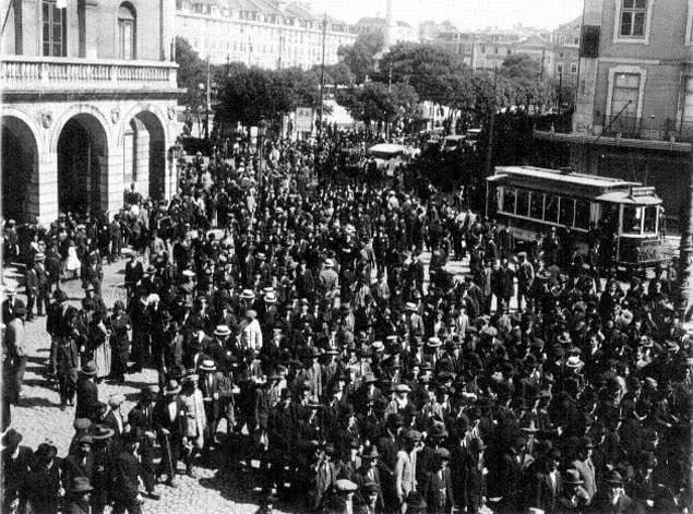 A dense crowd of men in hats and dark coats fills a street beside an arcaded building, with an electric tram behind them and trees and tall houses in the distance.
