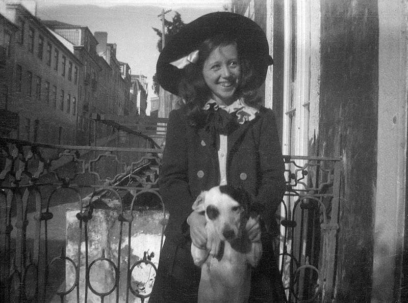 A smiling girl in a wide-brimmed hat and a dark buttoned coat stands on a balcony with a wrought-iron railing, holding up a small white dog with dark ears. A street of tall houses runs away behind her.