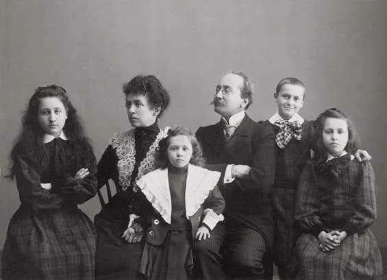 A studio portrait of a family of six. A seated woman in a dark dress with a lace collar and a man with spectacles and a moustache sit among four girls in dark dresses, one of them small and wearing a wide white collar.