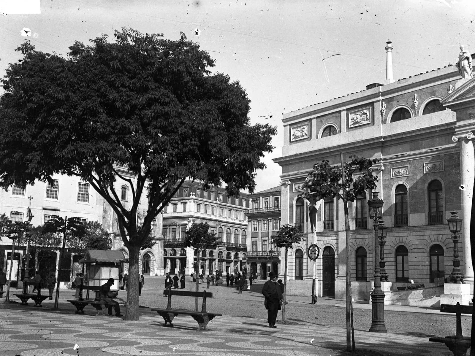 A black and white street scene. On the right rises the side of a large neoclassical building with tall arched windows, carved panels and a statue on the corner of the roof. On the left a broad tree shades benches and a kiosk on a pavement of wave-patterned mosaic, and men in hats walk towards a square lined with tall buildings, one with a Gothic-style arched doorway.