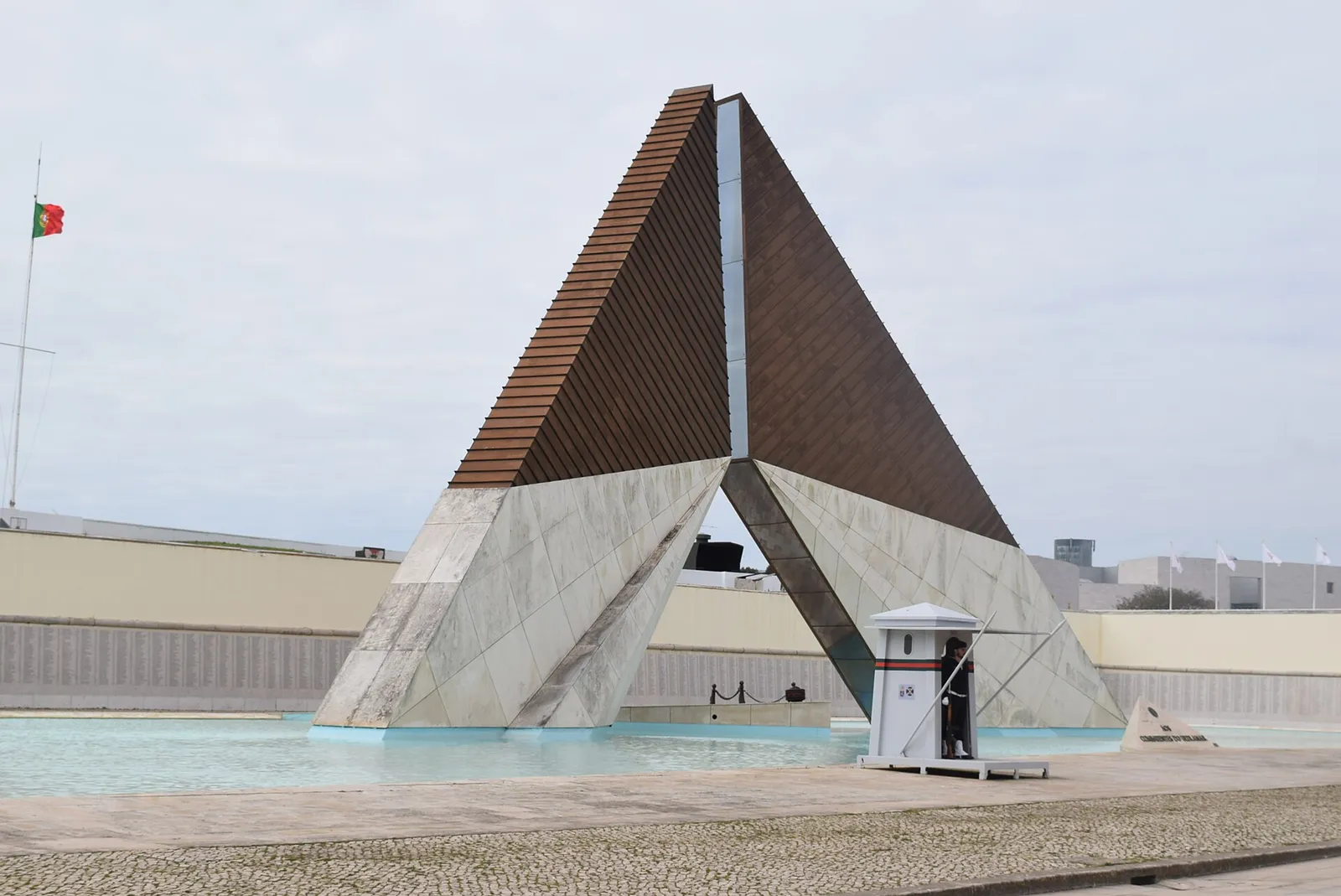 A tall triangular monument, dark brown above and pale stone below, rises from a shallow pool, with a sentry box and a guard beside it and a Portuguese flag on a pole to the left.