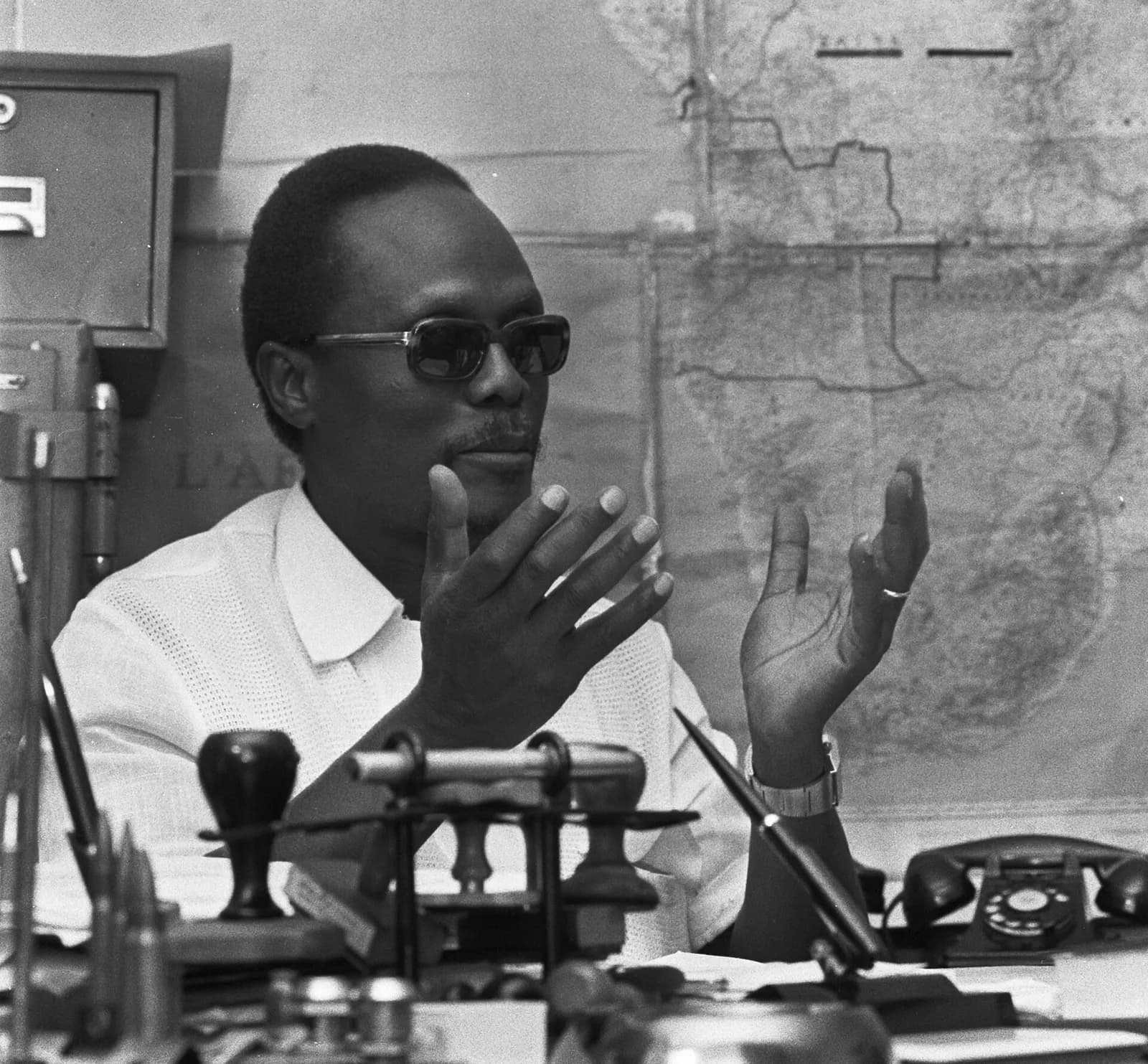 A man in dark glasses and a white shirt sits at a cluttered desk, gesturing with both hands. A large map of Africa is pinned to the wall behind him.