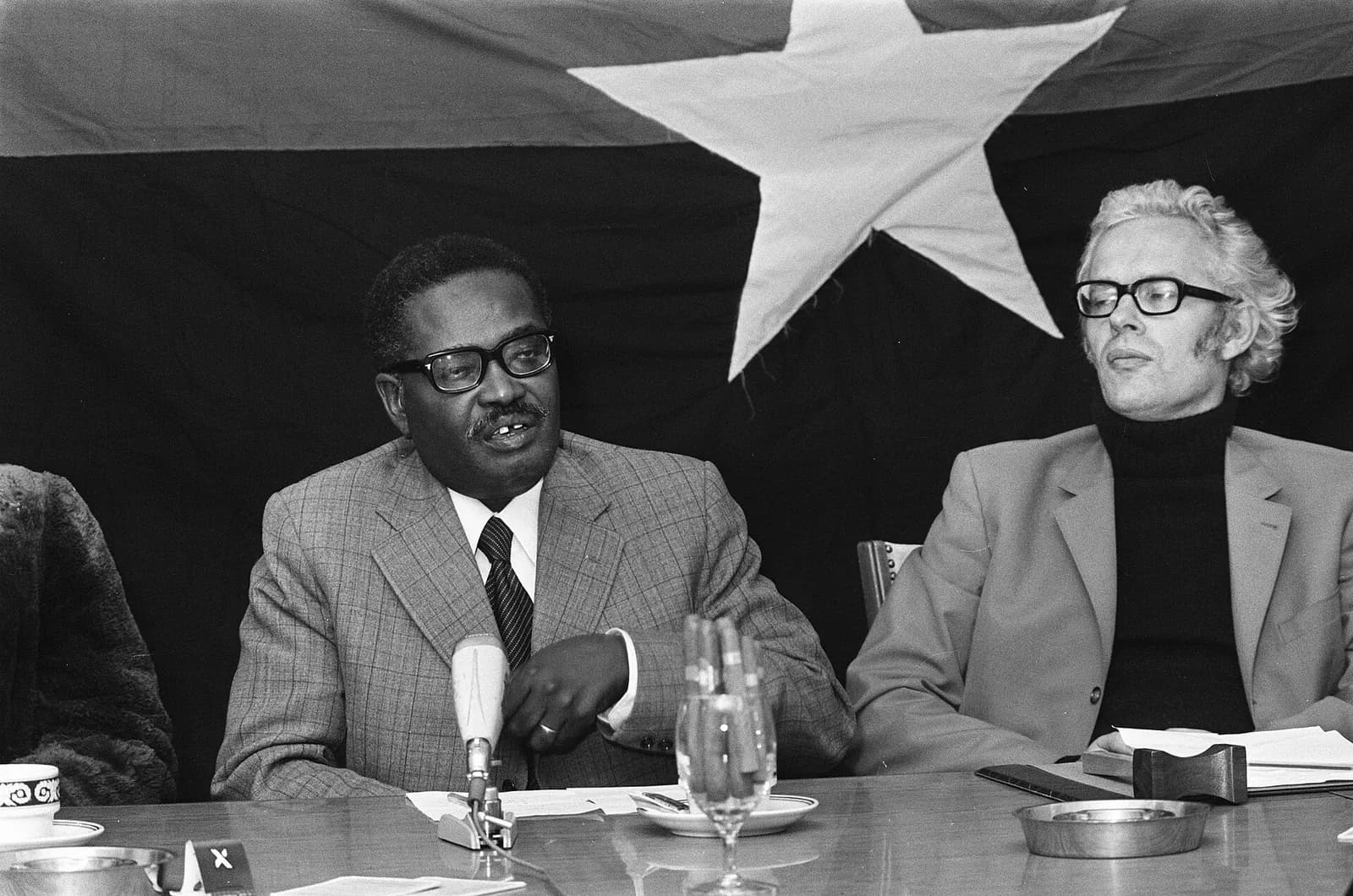 A man in glasses, a checked suit and a tie speaks at a table with a microphone in front of him. A fair-haired man in a polo-neck sits beside him, and a large flag with a big star hangs on the wall behind them.