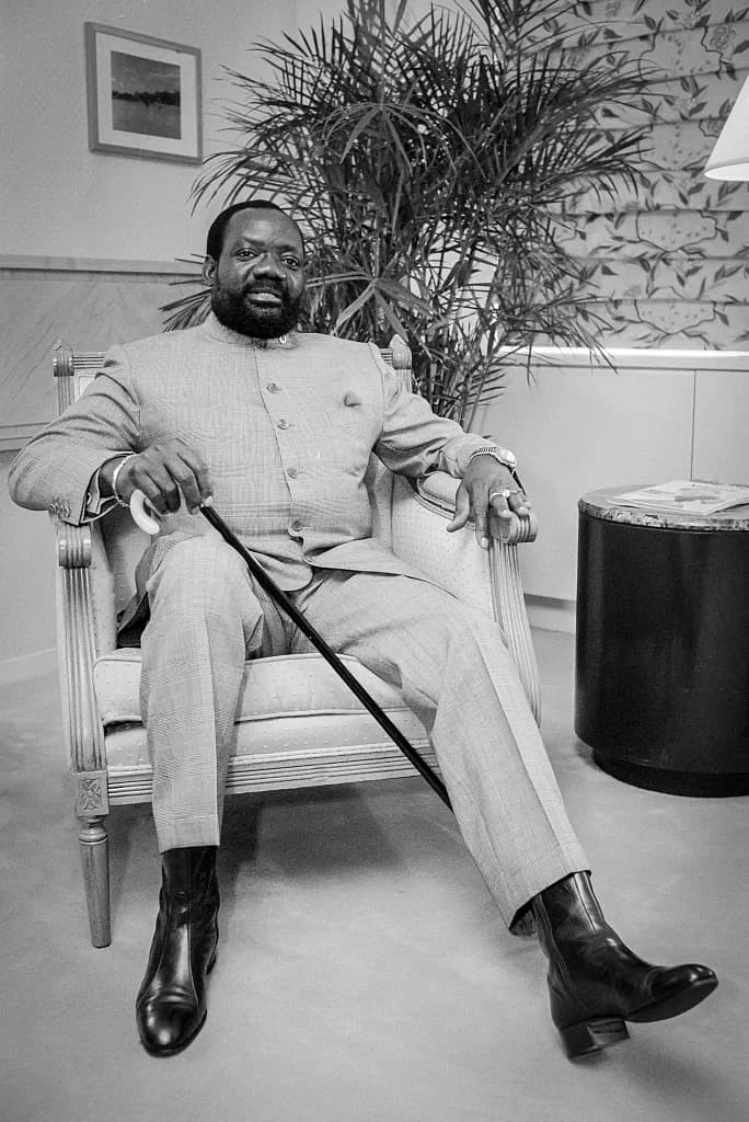 A bearded man in a light buttoned tunic and trousers sits in an armchair holding a walking cane, in front of a potted palm.