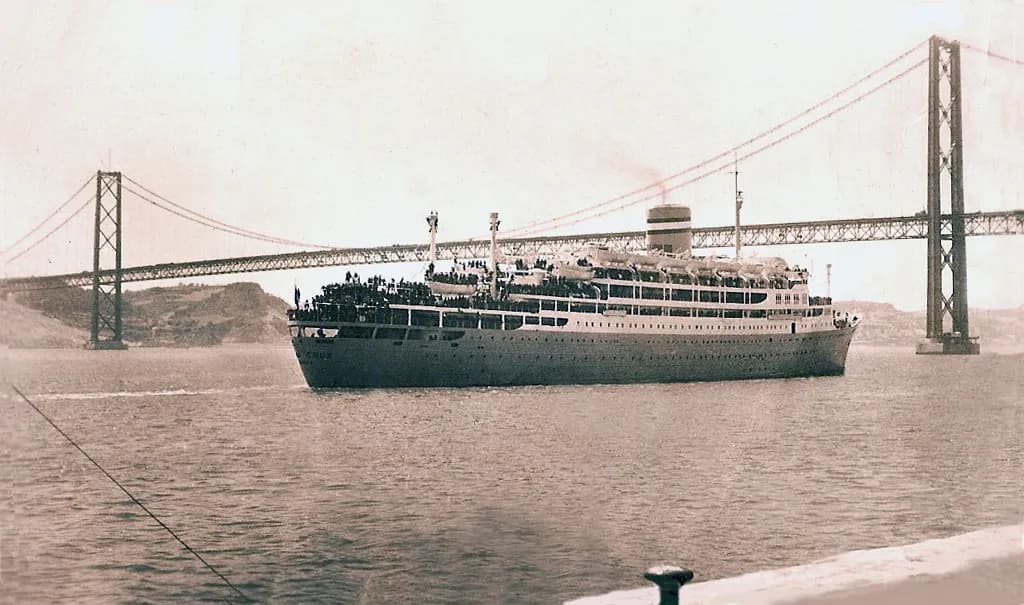 A large white ocean liner, its decks crowded with people, sails down a wide river under a long suspension bridge with tall towers.