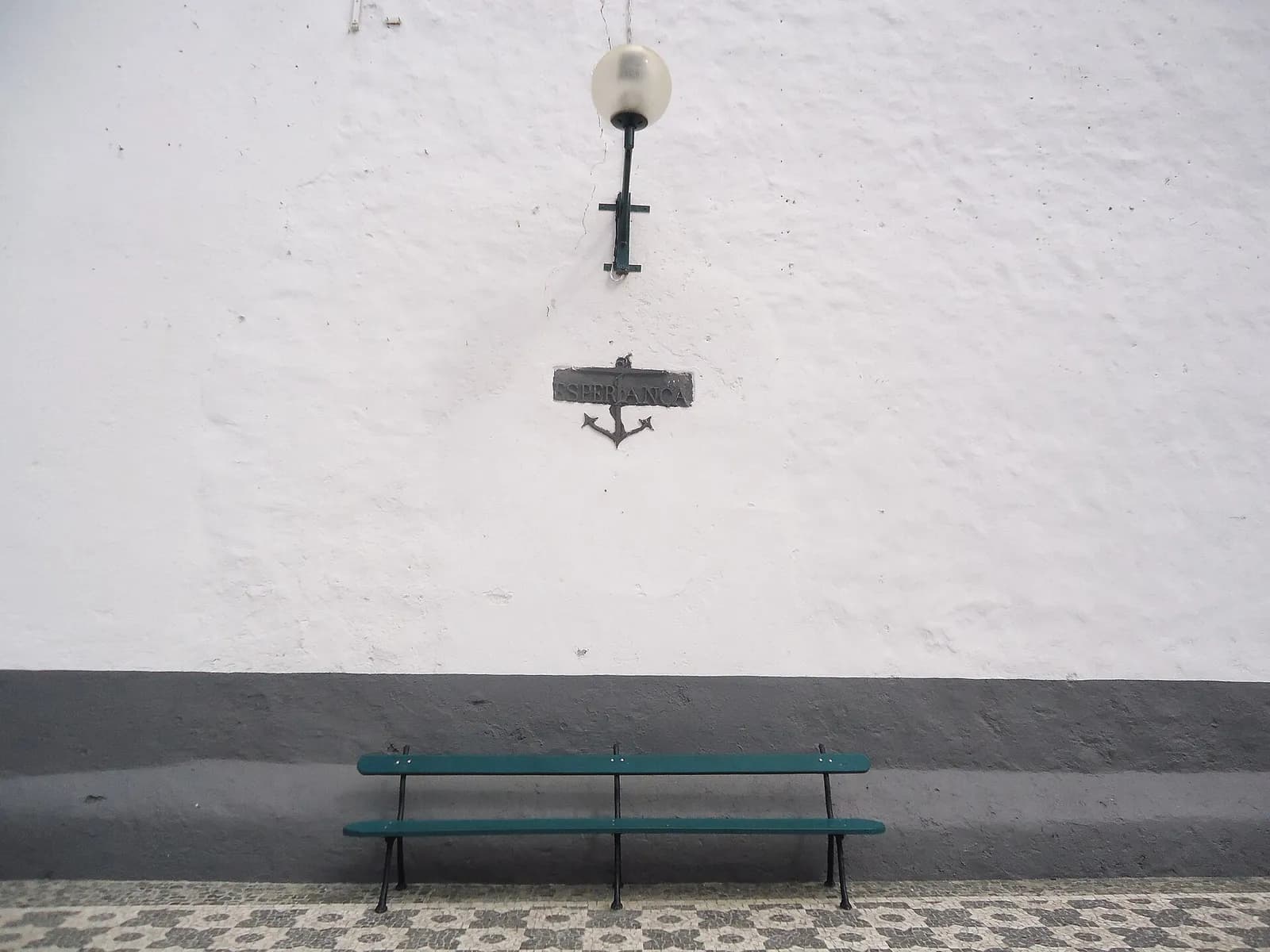 A green wooden bench against a whitewashed wall with a grey base, on black and white cobbles. Above it a small dark plaque reading Esperança, with an anchor beneath, hangs below a wall lamp.