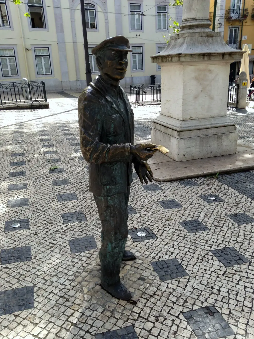Bronze statue of a man in a peaked cap and jacket standing on a cobbled square, holding out a lottery ticket in one hand, with a stone column base and buildings behind him.