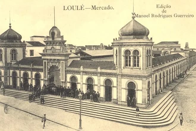 Sepia postcard of a long market building with arched doorways and windows and onion-shaped domes on its corner towers, reached by a broad flight of steps on which groups of men in hats stand talking.