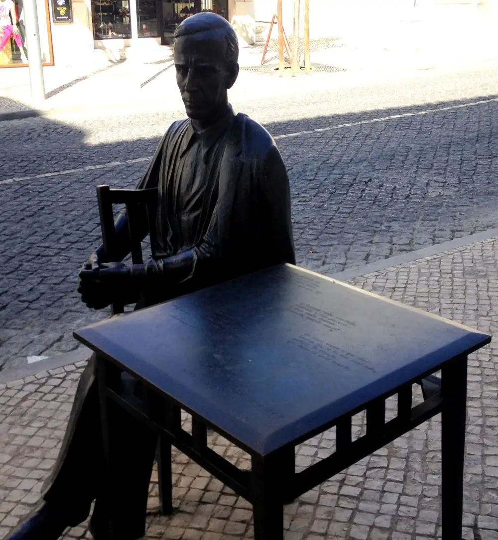 Bronze statue of a thin, short-haired man in a jacket sitting on a wooden-style chair with his hands folded, behind a square bronze table whose top is engraved with lines of verse, on a cobbled pavement.
