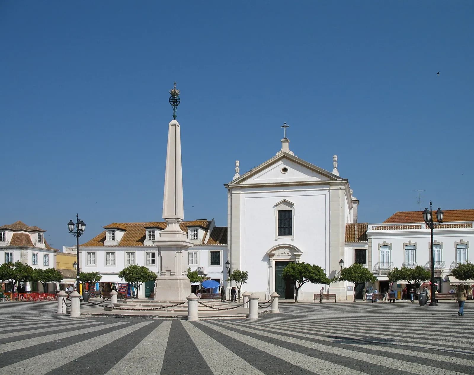 A wide square paved in black and white stripes under a clear blue sky, with a tall white obelisk left of centre and a white church with a triangular pediment and a cross at the far side, flanked by low two-storey buildings and orange trees.