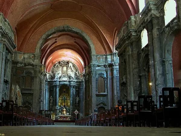 The nave of a large church with tall, scarred and blackened stone pilasters, a gilded altar at the far end under a reddish vault and rows of wooden chairs.