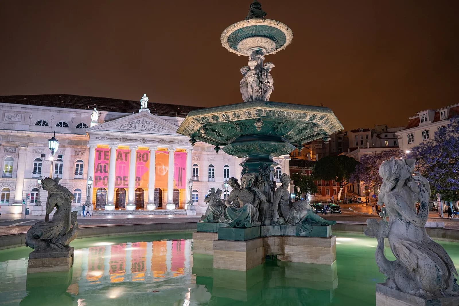 The neoclassical columned portico of the Teatro Nacional D. Maria II lit at night, seen behind a large bronze fountain with sculpted figures in the Rossio.