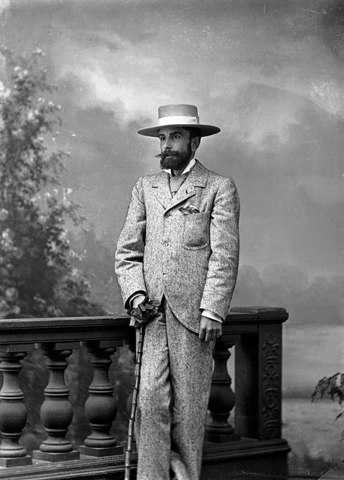 Black-and-white studio portrait of a bearded man in a light flecked tweed suit and a straw boater, standing beside a carved balustrade in front of a painted backdrop of sky and foliage, holding a cane and gloves.