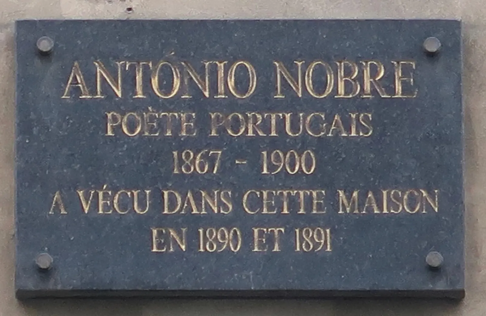 A dark grey stone plaque fixed to a wall with four bolts, inscribed in gold capital letters in French.