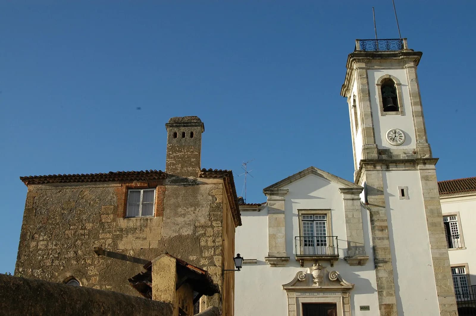 A weathered stone building with a tiled roof, a single window and a tall chimney stands on the left against a deep blue sky. To its right is a white church front with stone trim and a tall white bell tower with a clock.