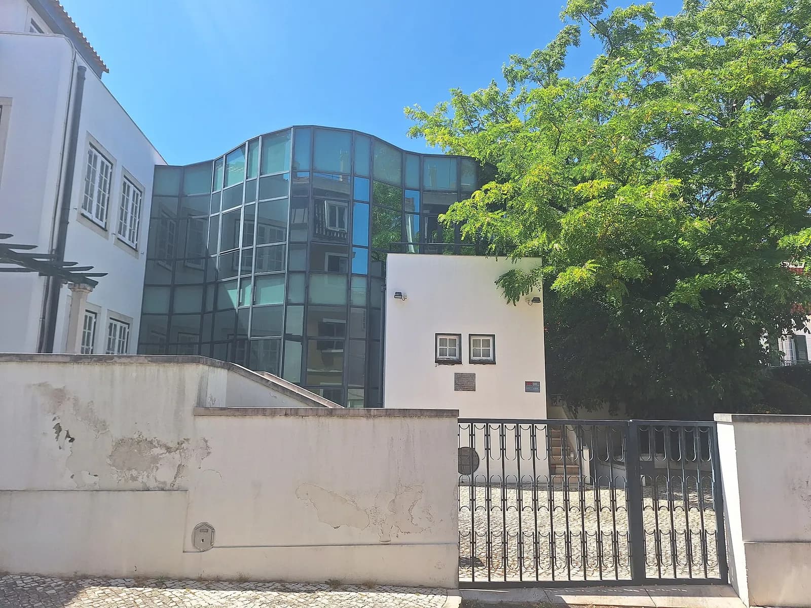 A small white building with two square windows and two plaques stands behind a whitewashed wall and an iron gate, joined to a curved, multi-storey wing of dark glass. A leafy tree fills the right side and a white town house the left, under a clear blue sky.