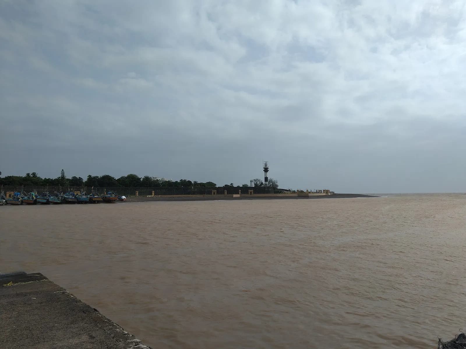 A wide brown river mouth under a grey, cloudy sky. On the far bank, beyond a row of moored fishing boats, a low fortified wall runs along the shore to a black-and-white lighthouse, with trees behind and the open sea to the right.