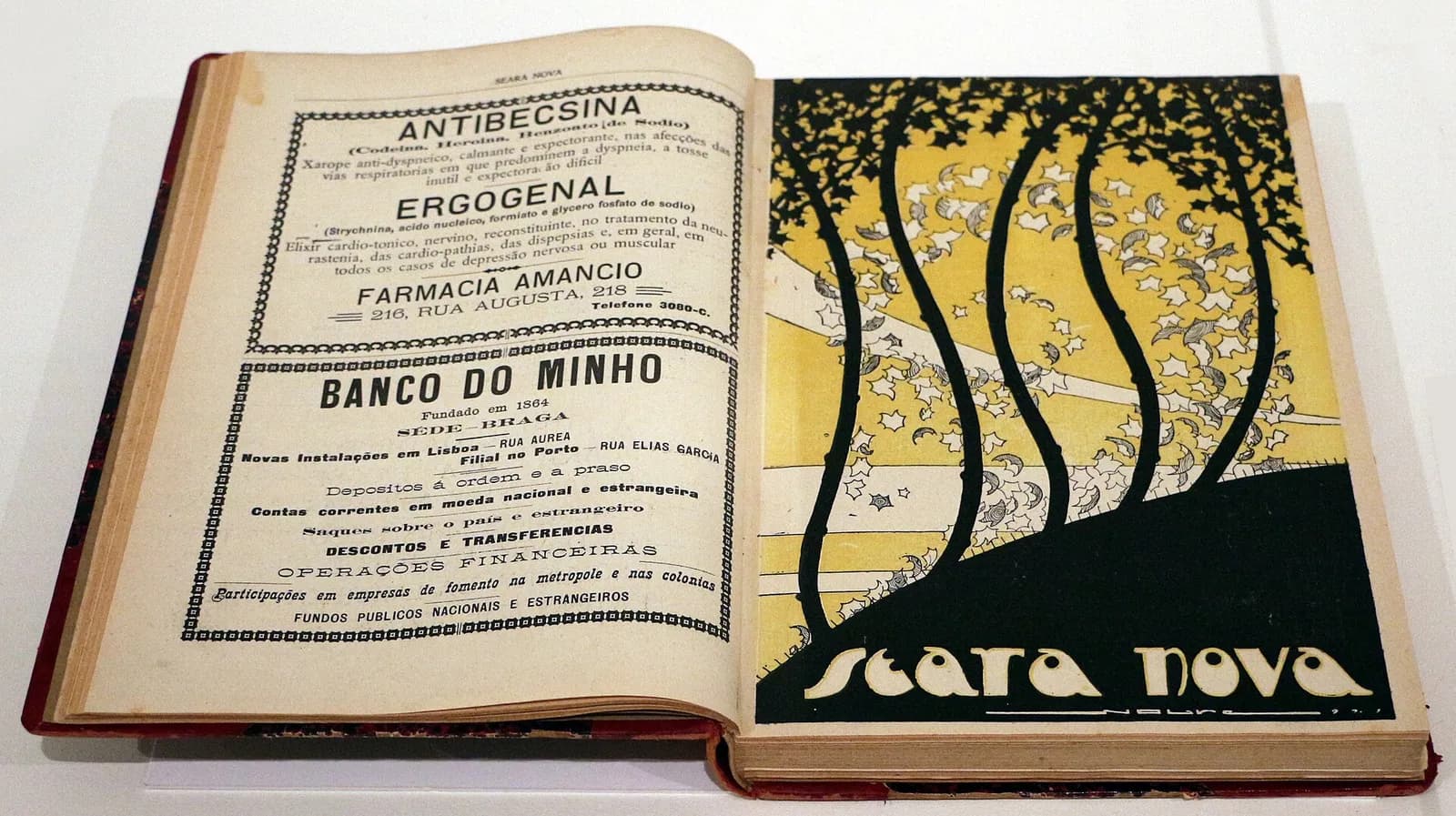 A thick bound volume lies open in a museum case. The left page is filled with old advertisements for medicines, a Lisbon pharmacy and the Banco do Minho; the right page is a cover in black and yellow showing slender dark trees on a hill against a yellow sky full of falling leaves, with the title Seara Nova in large stylised letters.