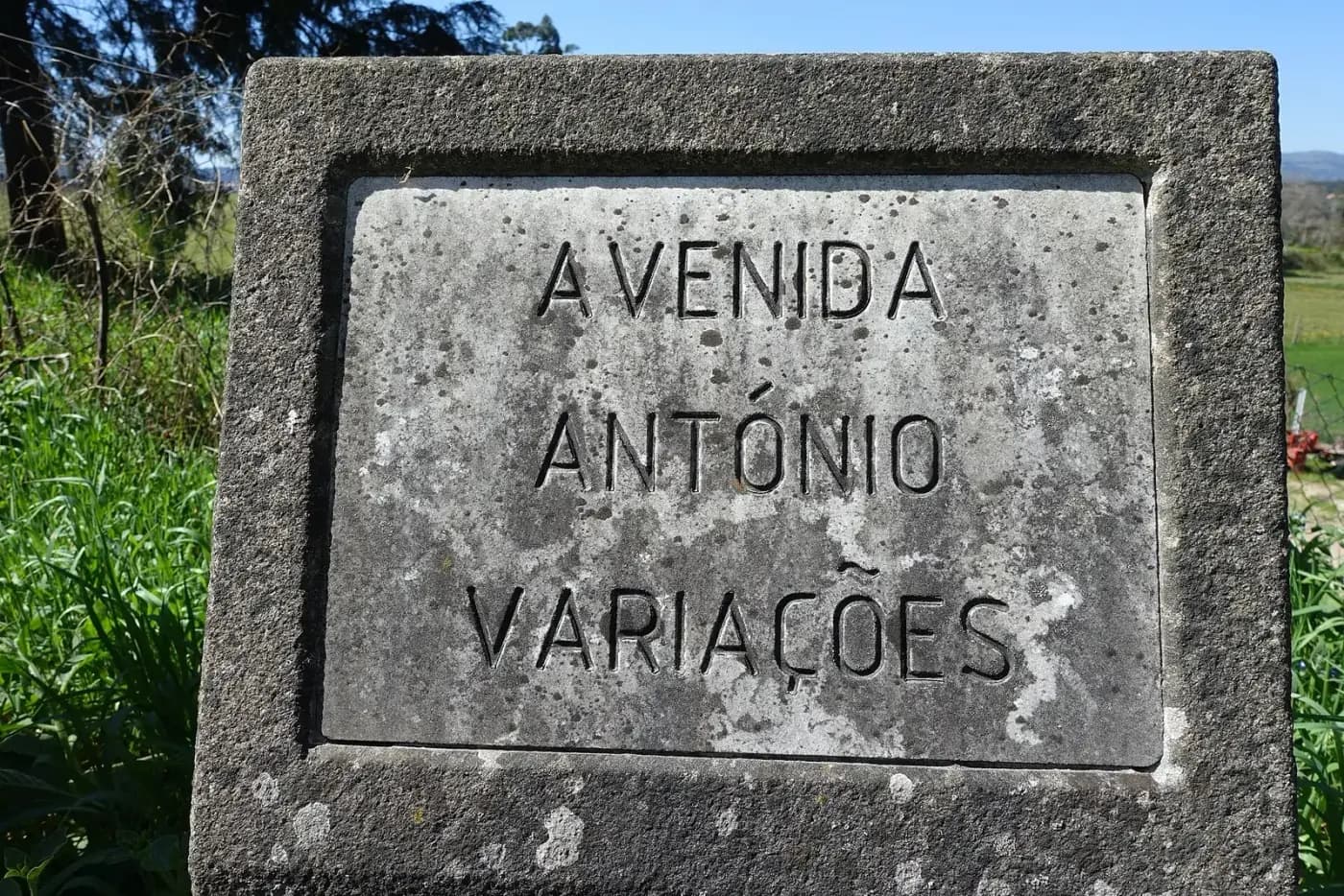 A weathered stone street sign in a thick stone frame, carved with the words Avenida António Variações, with long grass, trees and fields behind.
